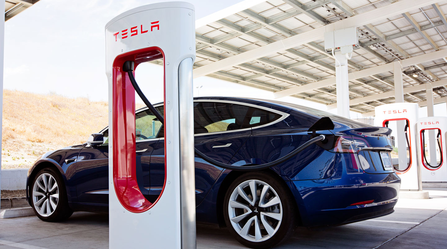 Tesla Model 3 grise en recharge sur une borne Superchargeur en périphérie urbaine, câble branché, écran de la borne affichant l'état de recharge en français, lumière dorée de fin d'après-midi